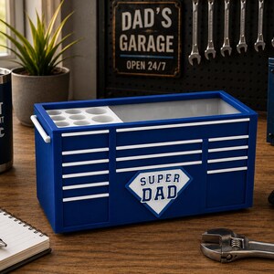 May include: A blue toolbox with white stripes and the words "SUPER DAD" on the front. A blue tumbler with the words "BUILT NOT BOUGHT" is on the left. A wrench and gloves are on the right.