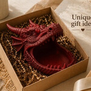 May include: A red dragon-shaped decorative bowl with an open mouth, presented in a brown gift box with shredded paper. The box is next to a small gift box with a bow. The text "Unique gift idea" is visible.