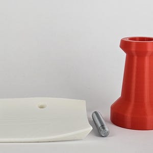 May include: A red, 3D-printed object with a wide base and a narrow top, resembling a pushpin. A white, curved, square object with a hole and a small, silver cylinder are also present. The objects are on a white surface.