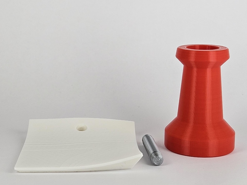 May include: A red, 3D-printed object with a wide base and a narrow top, resembling a pushpin. A white, curved, square object with a hole and a small, silver cylinder are also present. The objects are on a white surface.