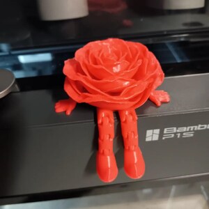 May include: A 3D printed red rose figurine with legs and arms. The rose is sitting on a black surface with the text "Bambi P1S" printed on it.