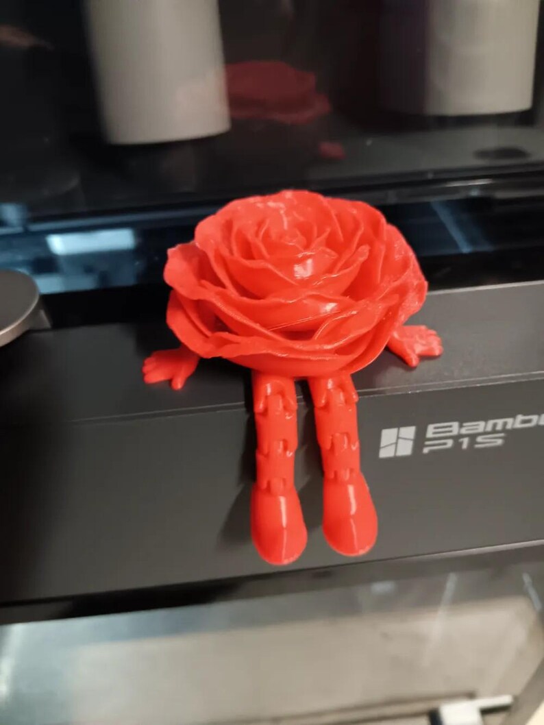 May include: A 3D printed red rose figurine with legs and arms. The rose is sitting on a black surface with the text "Bambi P1S" printed on it.