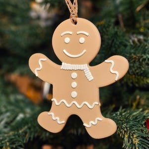 May include: A tan gingerbread man Christmas ornament with white icing details, including a scarf, buttons, and wavy lines. The ornament is hanging in front of a blurred green Christmas tree.