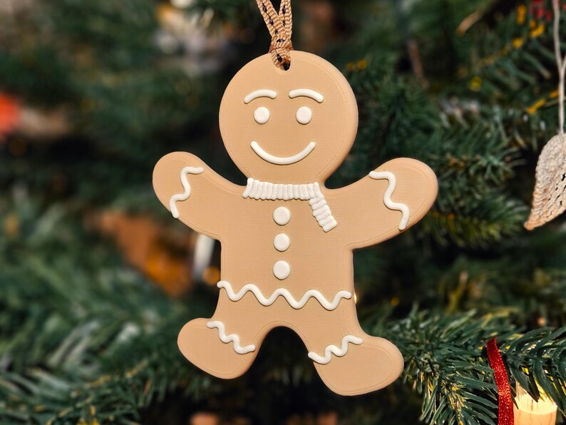 May include: A tan gingerbread man Christmas ornament with white icing details, including a scarf, buttons, and wavy lines. The ornament is hanging in front of a blurred green Christmas tree.