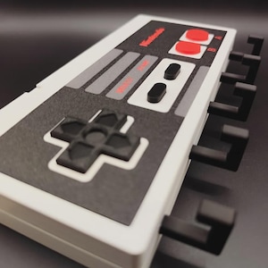 May include: A white and grey wall hook in the shape of a Nintendo controller, featuring black hooks. The controller design includes red buttons and the Nintendo logo, resembling the classic NES controller.