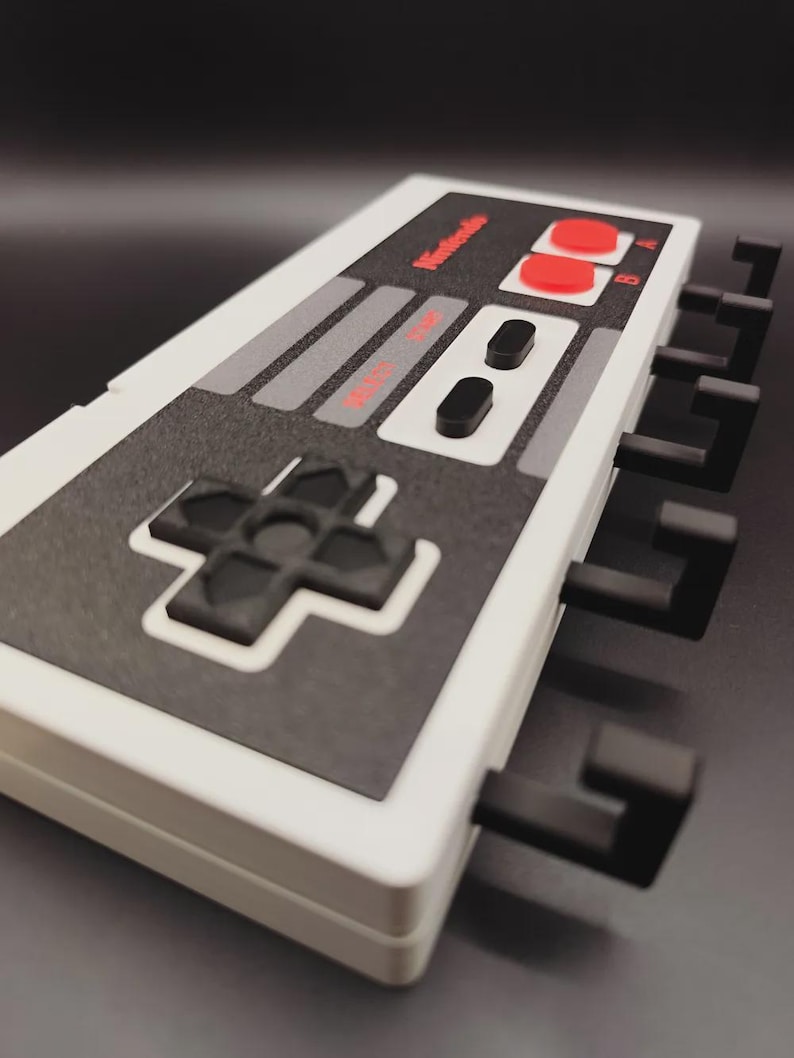May include: A white and grey wall hook in the shape of a Nintendo controller, featuring black hooks. The controller design includes red buttons and the Nintendo logo, resembling the classic NES controller.