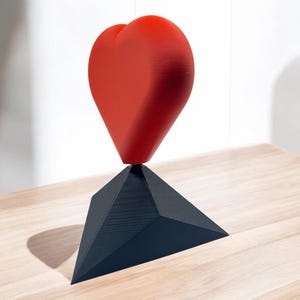 Pu&ograve; includere: Una scultura a forma di cuore rosso in equilibrio su una base triangolare nera. La scultura &egrave; realizzata in un materiale liscio e lucido e si trova su una superficie in legno chiaro.
