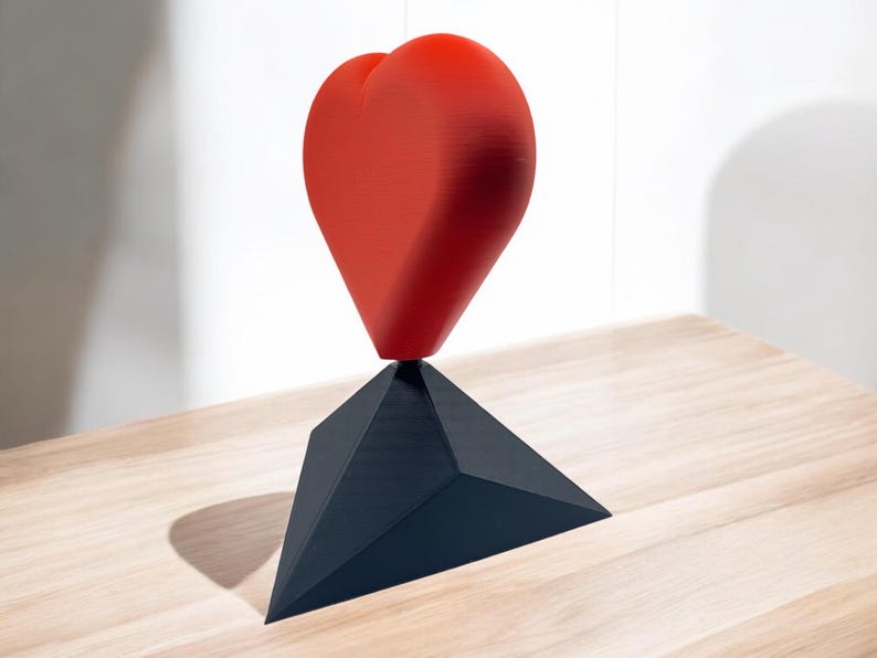 Pu&ograve; includere: Una scultura a forma di cuore rosso in equilibrio su una base triangolare nera. La scultura &egrave; realizzata in un materiale liscio e lucido e si trova su una superficie in legno chiaro.