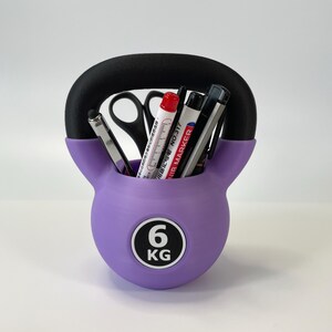 May include: A purple kettlebell-shaped desk organizer with a black handle. The organizer holds pens, markers, and scissors. The front of the organizer has a white circle with the number "6" and the letters "KG".