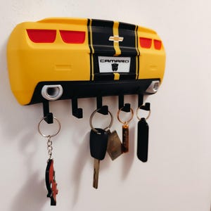 Puede incluir: Llavero amarillo con forma de Camaro con rayas negras y la palabra "CAMARO" en la matr&iacute;cula. El llavero tiene seis ganchos negros para colgar llaves. Varias llaves y llaveros cuelgan de los ganchos.