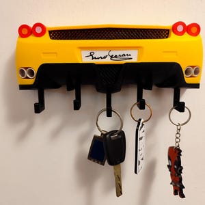 Puede incluir: Llavero con forma de coche amarillo y negro con cuatro ganchos. El llavero tiene un dise&ntilde;o trasero de coche con luces traseras y tubos de escape. Varias llaves y llaveros cuelgan de los ganchos. El llavero est&aacute; montado en una pared blanca.