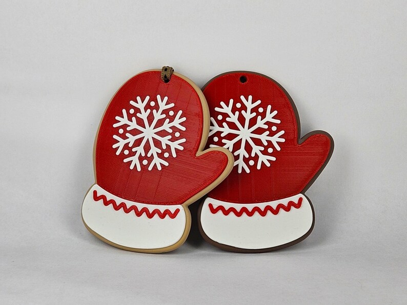 K&ouml;nnte beinhalten: Zwei rote, handschuhf&ouml;rmige Weihnachtsschmuckst&uuml;cke mit wei&szlig;en Schneeflockenmustern. Jeder Handschuh hat eine wei&szlig;e Manschette mit einem roten Zickzackmuster. Die Ornamente bestehen aus einem strukturierten Material und haben oben ein Loch zum Aufh&auml;ngen.