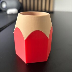 May include: A red and beige pencil-shaped desk organizer. The organizer is made of plastic and has a hexagonal base.