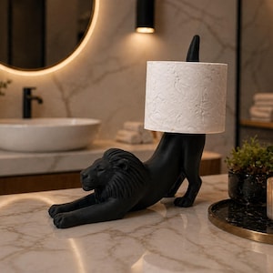 Pode incluir: Um porta-papel higi&ecirc;nico em forma de le&atilde;o preto. O le&atilde;o est&aacute; em uma pose de alongamento, sustentando um rolo de papel higi&ecirc;nico branco estampado. A decora&ccedil;&atilde;o do banheiro inclui um espelho redondo, uma pia branca e uma vela.