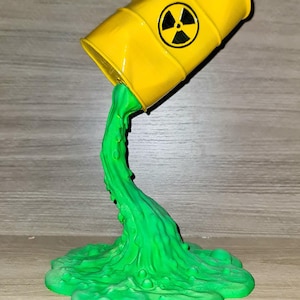 May include: A yellow barrel with a black radiation symbol is tilted, with green liquid pouring out. The liquid forms a puddle at the base. The barrel is a novelty item.