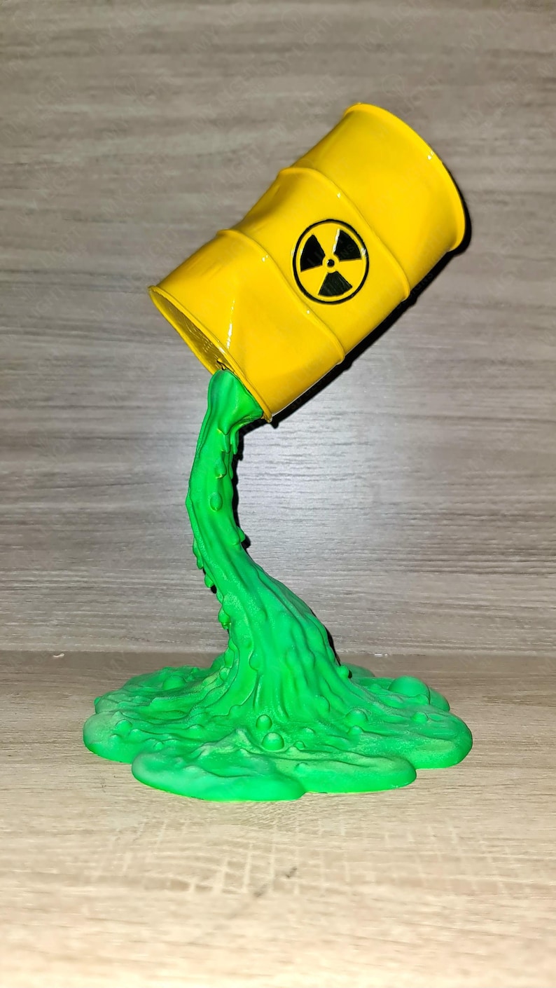 May include: A yellow barrel with a black radiation symbol is tilted, with green liquid pouring out. The liquid forms a puddle at the base. The barrel is a novelty item.