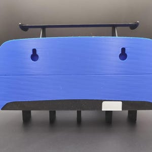 May include: A blue wall-mounted shelf with a black spoiler and black supports. The shelf has two circular holes and a black foam base. The shelf is designed to hold items and is suitable for various rooms.