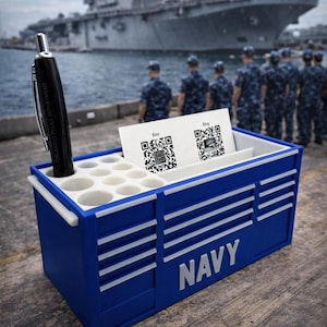 May include: A blue desk organizer with white accents and the word "NAVY" in white. The organizer holds a black pen and business cards with QR codes. A military ship and uniformed personnel are in the background.