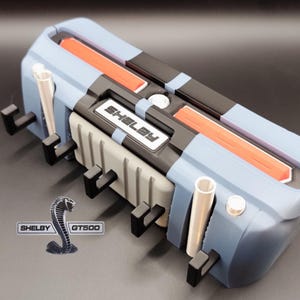 K&ouml;nnte beinhalten: Ein blaues, schwarzes und graues Modell eines Automotors mit dem Text "Shelby" auf der Vorderseite. Das Modell hat orangefarbene Akzente und ein silbernes Shelby GT500-Logo.