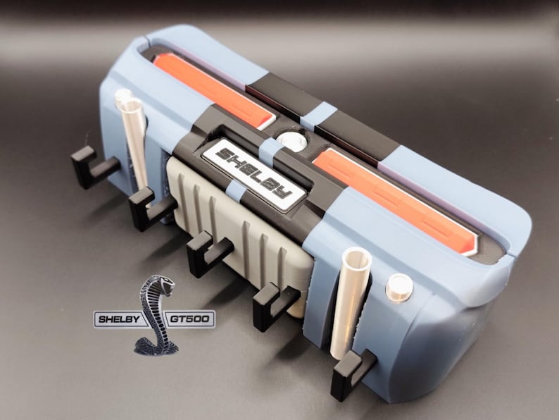 K&ouml;nnte beinhalten: Ein blaues, schwarzes und graues Modell eines Automotors mit dem Text "Shelby" auf der Vorderseite. Das Modell hat orangefarbene Akzente und ein silbernes Shelby GT500-Logo.