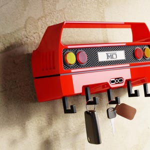 May include: A red car-shaped key holder with black hooks. The key holder has a handle and features yellow and red lights, a black grill, and the number "140". Keys hang from the hooks.