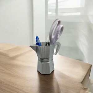 May include: A gray, 3D-printed desk organizer shaped like a stovetop espresso maker. It holds a blue pen, a black and white marker, and a pair of lavender scissors. The organizer sits on a wooden surface.