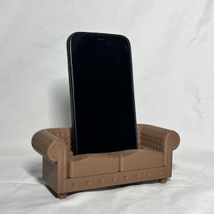 May include: A brown miniature sofa with a black smartphone standing upright in the middle of the sofa.