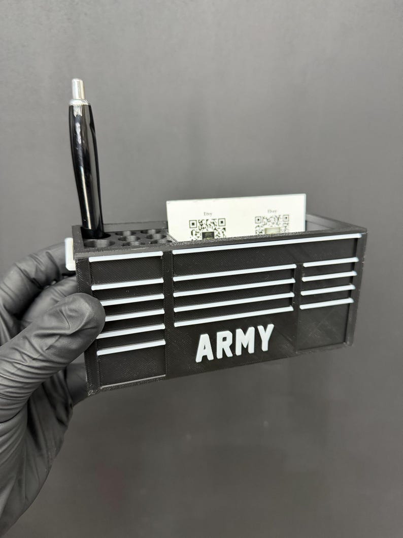 May include: Black desk organizer with the word "ARMY" in white block letters. The organizer has a slot for business cards and a pen holder with a black pen. The design includes horizontal white stripes.
