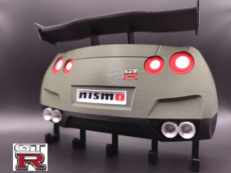 Może przedstawiać: Tył Nissana GT-R w kolorze oliwkowym, wydrukowany w 3D, z czarnym spojlerem i rurami wydechowymi. Posiada czerwone światła tylne, logo "GT-R" i emblemat "NISMO". Zawiera haczyki do wieszania.