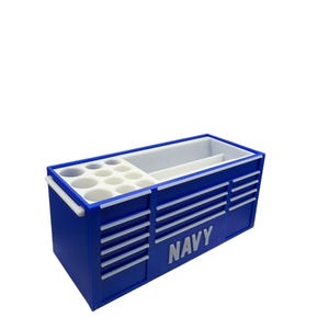 Op de afbeelding: Een blauwe bureau-organizer met het woord "NAVY" in het wit. De organizer heeft meerdere compartimenten, waaronder een sectie met ronde gaten en een groter open gebied. Het heeft witte accenten en handgrepen.