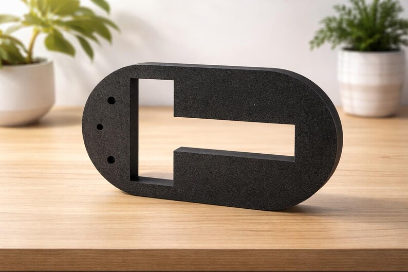 Op de afbeelding: Een zwarte, abstracte sculptuur met een ovale vorm en rechthoekige uitsparingen. Er zijn drie kleine gaten in de zijkant geboord. De sculptuur is gemaakt van een materiaal met textuur en staat op een houten oppervlak.