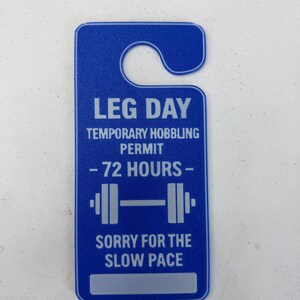K&ouml;nnte beinhalten: Blauer T&uuml;rh&auml;nger mit wei&szlig;em Text. Der Text lautet "LEG DAY TEMPORARY HOBBLING PERMIT - 72 HOURS -" und "SORRY FOR THE SLOW PACE." Eine Hantel-Grafik befindet sich in der Mitte.