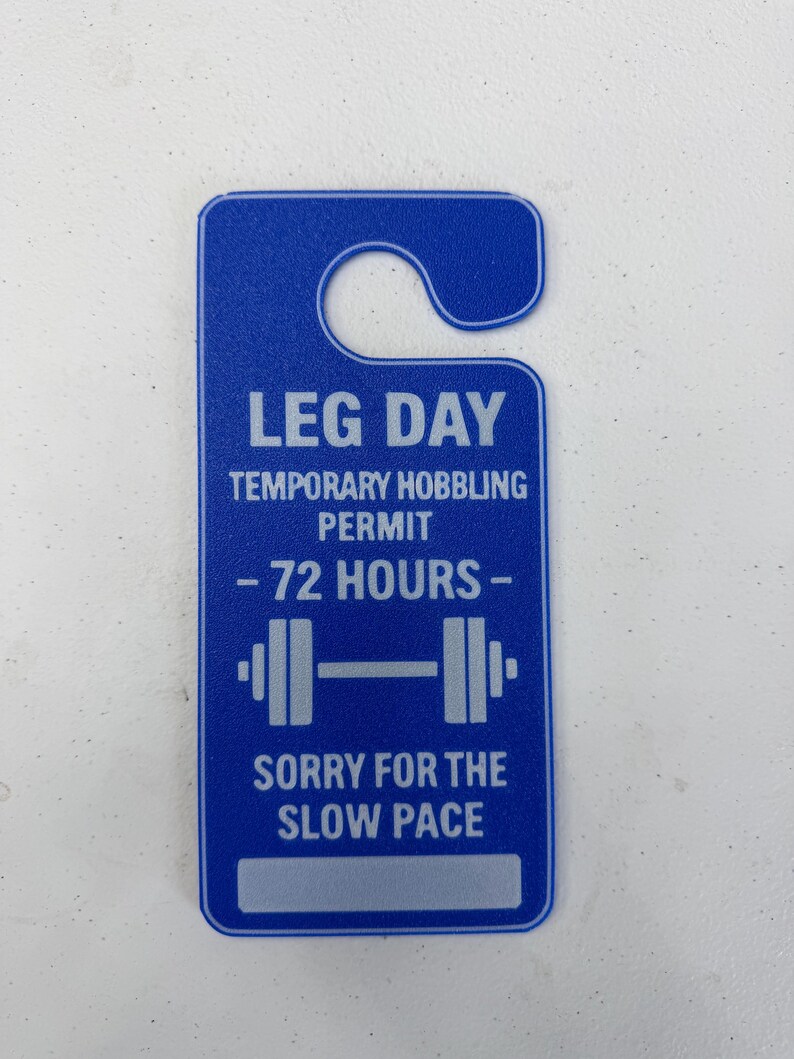 K&ouml;nnte beinhalten: Blauer T&uuml;rh&auml;nger mit wei&szlig;em Text. Der Text lautet "LEG DAY TEMPORARY HOBBLING PERMIT - 72 HOURS -" und "SORRY FOR THE SLOW PACE." Eine Hantel-Grafik befindet sich in der Mitte.