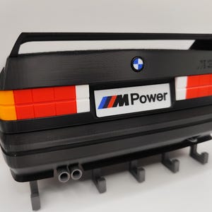 May include: Black decorative wall mount featuring the rear of a BMW M3 car. The design includes the BMW logo, "M Power" text, and taillights with orange, red, and white sections. Includes hooks for hanging items.