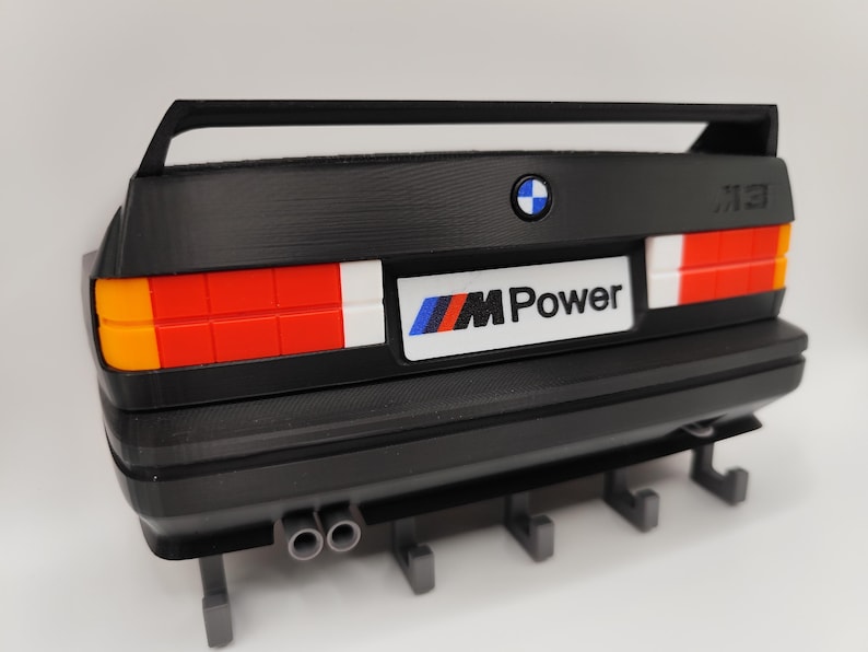 May include: Black decorative wall mount featuring the rear of a BMW M3 car. The design includes the BMW logo, "M Power" text, and taillights with orange, red, and white sections. Includes hooks for hanging items.