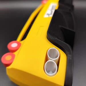 Puede incluir: Un coche de juguete amarillo y negro con detalles rojos y blancos. El coche de juguete tiene dos tubos de escape blancos y un peque&ntilde;o rect&aacute;ngulo blanco en el lateral. El coche tiene una forma redondeada y parece estar impreso en 3D.