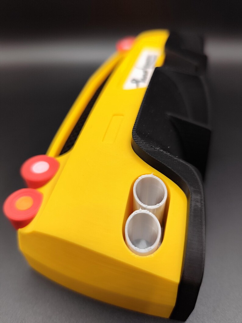 Puede incluir: Un coche de juguete amarillo y negro con detalles rojos y blancos. El coche de juguete tiene dos tubos de escape blancos y un peque&ntilde;o rect&aacute;ngulo blanco en el lateral. El coche tiene una forma redondeada y parece estar impreso en 3D.