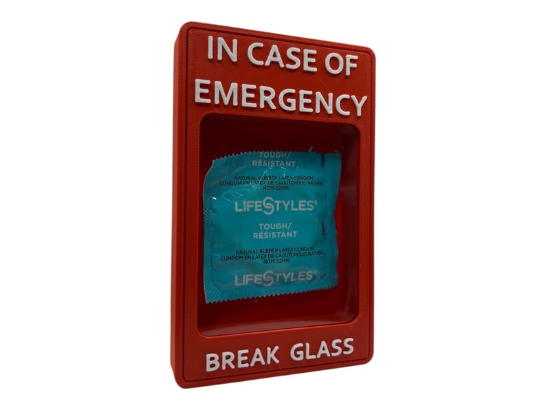 May include: A red plastic box with the words "IN CASE OF EMERGENCY" and "BREAK GLASS" in white lettering. Inside the box is a blue and white Lifestyles condom packet.