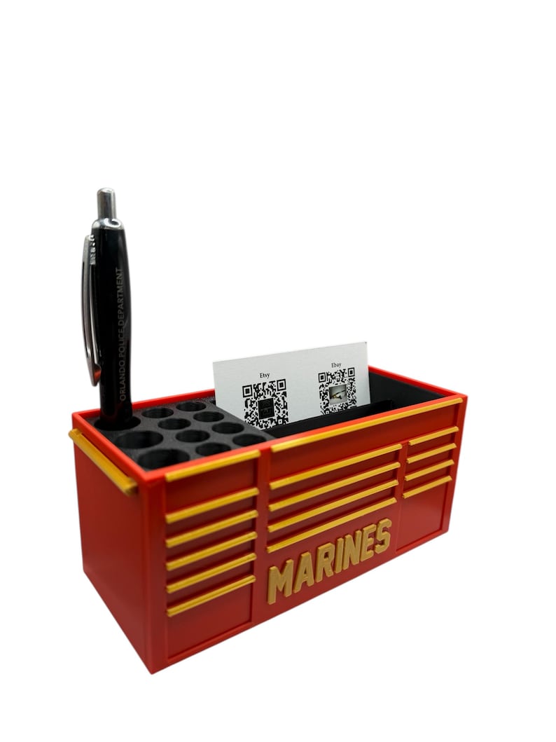 May include: A red and gold desk organizer with the word "MARINES" in gold lettering. The organizer has a section for pens, a slot for business cards, and a black pen. The pen has the text "ORLANDO POLICE DEPARTMENT".
