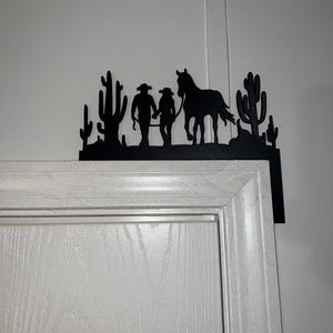 May include: Black metal silhouette of two cowboys and a horse walking through a desert landscape with cacti. The silhouette is mounted on a white door frame.