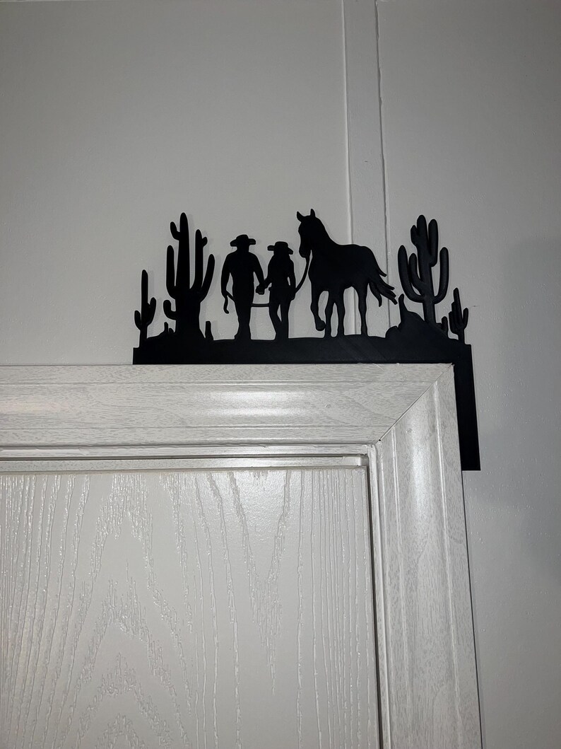 May include: Black metal silhouette of two cowboys and a horse walking through a desert landscape with cacti. The silhouette is mounted on a white door frame.