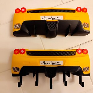 Puede incluir: Dos percheros de pared amarillos con forma de coche, detalles negros y el texto "Suno Ferrari". Cada perchero tiene luces traseras rojas y tubos de escape plateados. Los percheros est&aacute;n dise&ntilde;ados para parecerse a la parte trasera de un coche deportivo.