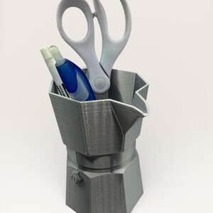 May include: A gray, 3D-printed desk organizer shaped like a stovetop espresso maker. It holds a pair of white scissors, a white marker, and a blue correction pen. The organizer is made of a textured plastic material.