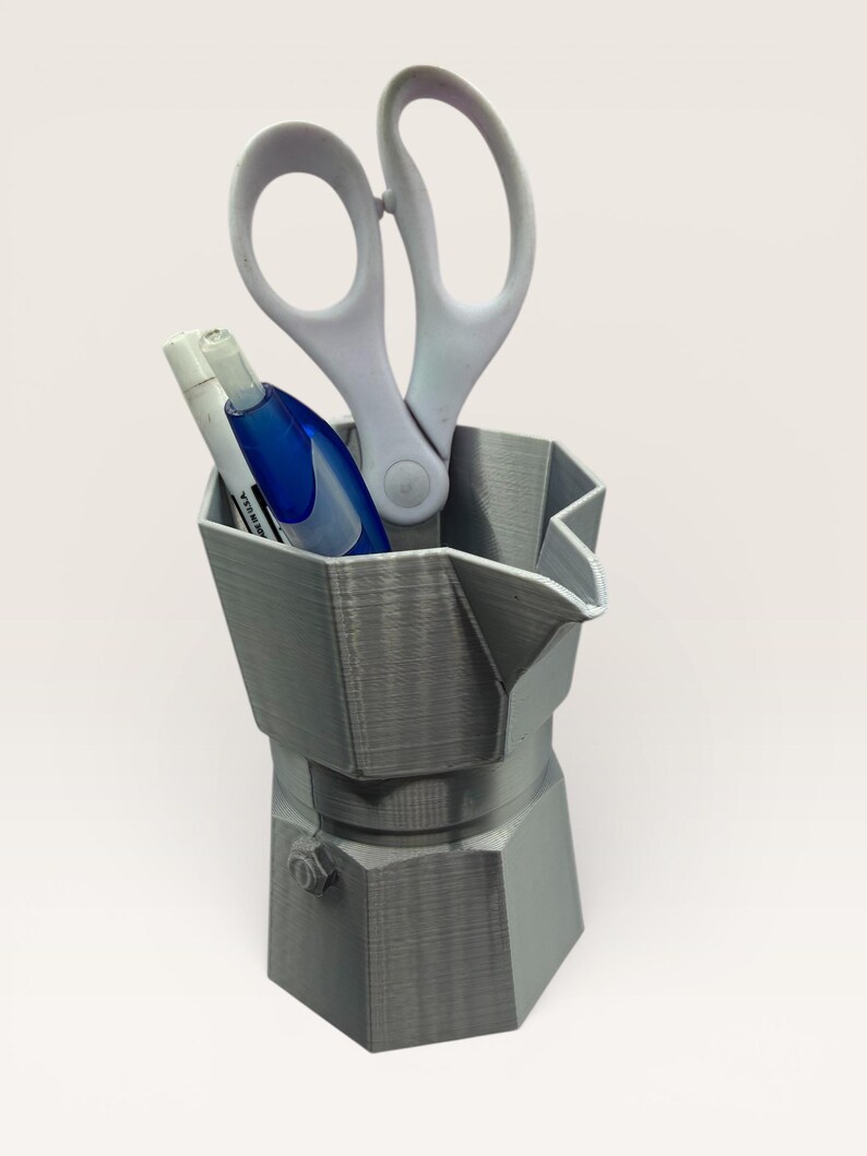 May include: A gray, 3D-printed desk organizer shaped like a stovetop espresso maker. It holds a pair of white scissors, a white marker, and a blue correction pen. The organizer is made of a textured plastic material.
