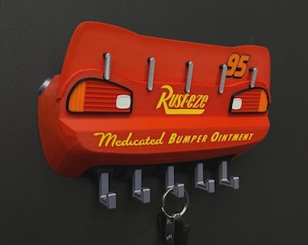 Puede incluir: Un llavero de pared rojo con forma de parte trasera de coche de carreras. Presenta el número 95, el logotipo de Rust-eze y el texto Medicated Bumper Ointment en amarillo. La unidad incluye cinco ganchos plateados, con un llavero metálico.