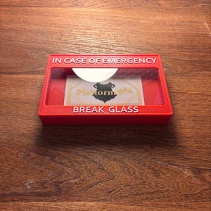 May include: A red break glass box with the text "IN CASE OF EMERGENCY" and "BREAK GLASS" in white lettering. The box has a clear plastic window with a card inside that says "Platform 9 3/4" with a black silhouette of a wizard's hat.