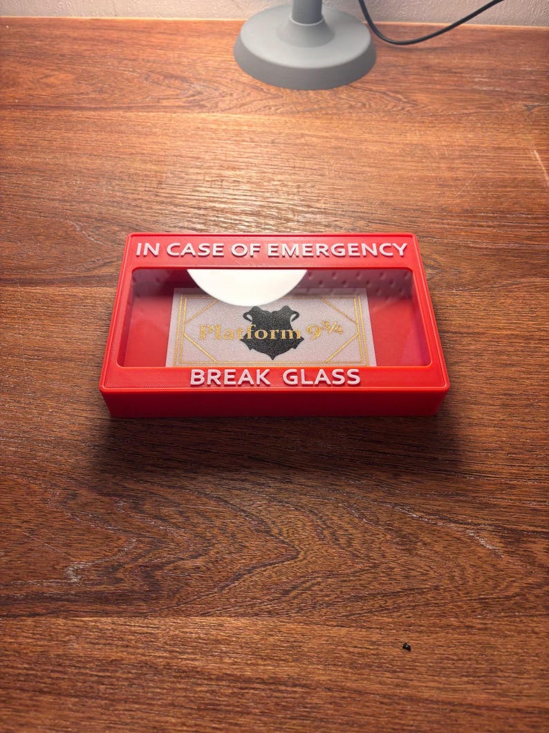 May include: A red break glass box with the text "IN CASE OF EMERGENCY" and "BREAK GLASS" in white lettering. The box has a clear plastic window with a card inside that says "Platform 9 3/4" with a black silhouette of a wizard's hat.