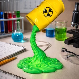 May include: A yellow barrel with a black radiation symbol is tilted, pouring out a stream of bright green liquid. The scene is set in a lab with beakers, test tubes, and a microscope, suggesting a science or novelty theme.