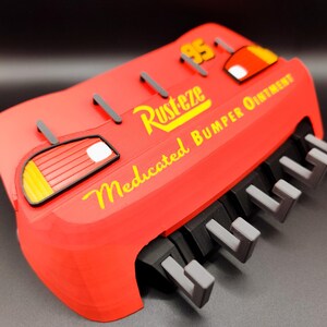 May include: A red, 3D-printed replica of a car bumper, featuring the text "Rusteze Medicated Bumper Ointment" in yellow. The design includes a "95" and car-like details. The bumper has gray and black accents.