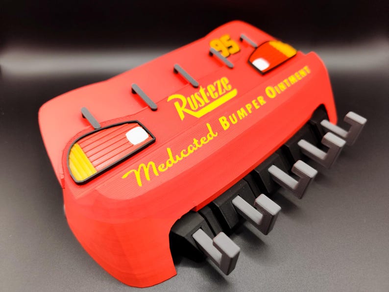 May include: A red, 3D-printed replica of a car bumper, featuring the text "Rusteze Medicated Bumper Ointment" in yellow. The design includes a "95" and car-like details. The bumper has gray and black accents.